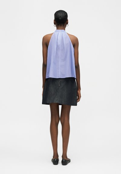 Femme aux cheveux courts, portant des boucles d'oreilles créoles, un haut léger bleu clair sans manches à col licou et une jupe en denim foncé, debout de dos avec des ballerines noires.