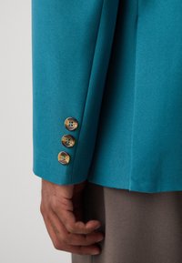 Türkise Blazerärmel mit drei goldenen Knöpfen und blauen Akzenten. Der Stoff hat eine glatte Textur; die Hand ruht auf grauem Stoff.