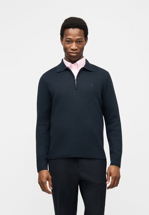 Homme portant un pull zippé bleu marine foncé sur une chemise à col rose clair, debout devant un fond clair uni.