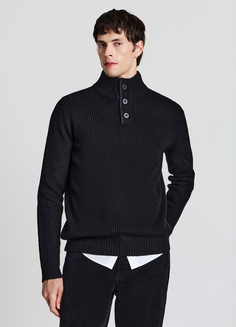 Jeune homme portant un pull noir en maille à col montant avec trois boutons, chemise blanche en dessous et pantalon noir, se tenant devant un fond uni.