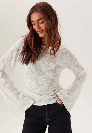 Femme aux longs cheveux bruns portant un haut en dentelle blanche à fleurs à manches longues évasées aux poignets et un pantalon noir, les mains dans les poches, regardant vers le bas en souriant.