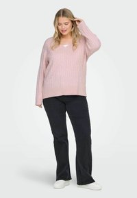 Roza pulover z pletenjem in V-izrezom ter dolgimi rokavi, v kombinaciji z temnimi širokimi jeans hlačami in belimi supergami. Mehka tekstura in sproščen kroj.