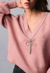 Svetlorožový pletený sveter s hlbokým V-výstrihom, so zúženými manžetami. Na pozadí visí strieborný náhrdelník s korálkami a malým príveskom vo forme mašle.