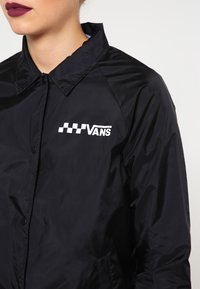 Henkilö, jolla on päällään musta valmentajantakki, jossa on valkoinen ruudullinen Vans-logo vasemmassa rinnassa, tumman viininpunainen huulipuna ja vaalea ihonväri.