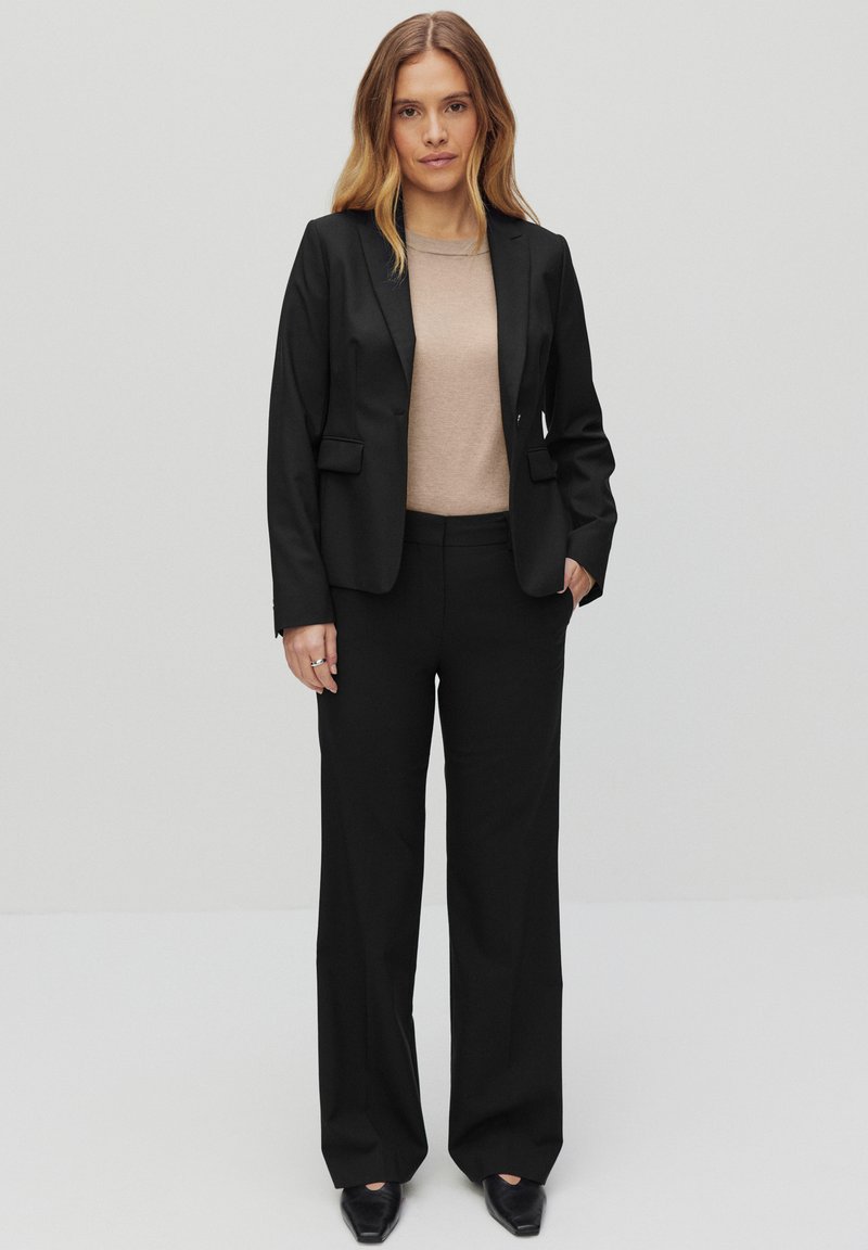 Svart kavaj med två framfickor, i kombination med en beige topp och svarta vida byxor. Outfiten kompletteras med eleganta svarta skor.