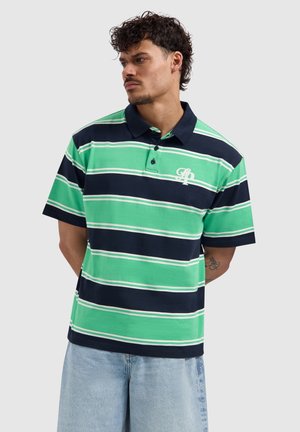 Man draagt een groen en marineblauw gestreepte polo met geborduurd logo, gecombineerd met lichtblauwe denimshorts, armen achter de rug.
