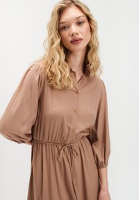 Blondynka z falującymi włosami ma na sobie brązową sukienkę z guzikami, z wciągniętą talią i rękawami 3/4 w stylu bufki, na białym tle.