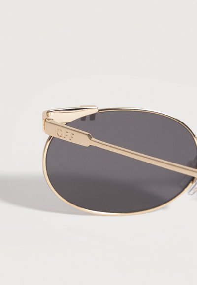 Lunettes de soleil ovales à monture dorée et verres foncés, posées sur une surface blanche, avec une gravure "OFF" sur la branche.
