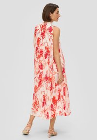 Femme portant une robe midi sans manches fluide avec un imprimé floral rouge, nouée au cou, associée à des sandales à talons nudes, se tenant de profil.