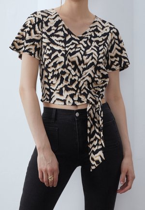Zwart- en crèmekleurige wikkeltop met korte mouwen, een V-hals en een strikdetail, voorzien van een getextureerd dierenprintpatroon. Gecombineerd met zwarte, high-waisted broeken.