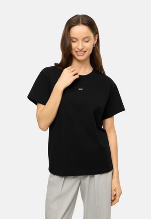 Vrouw met halflang bruin haar draagt een zwart T-shirt met klein logo en lichtgrijze broek, glimlachend en naar beneden kijkend.