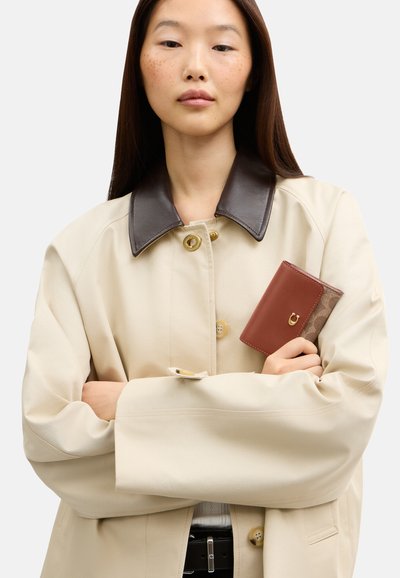 Femme portant un manteau beige avec un col foncé tenant un petit portefeuille en cuir marron sur un fond blanc uni.