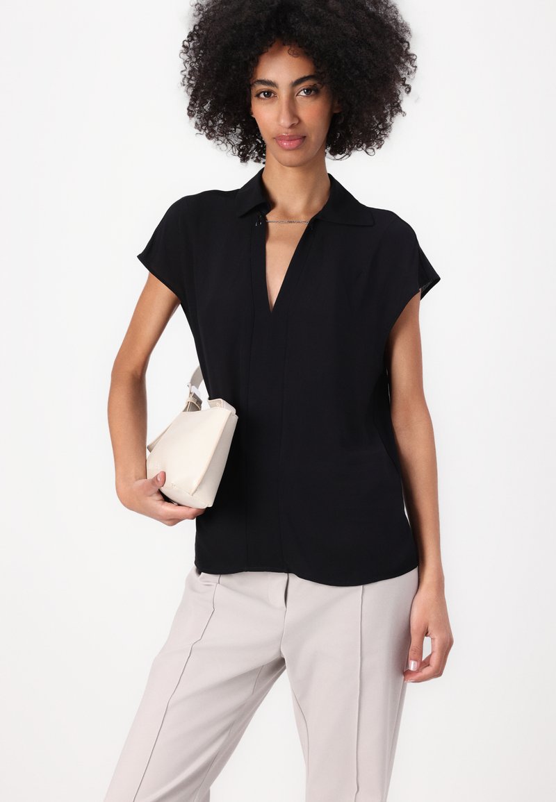 Blouse noire à manches courtes avec un décolleté en V, associée à un pantalon gris clair. Le modèle tient un petit sac à main beige.