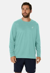 Camicia lunga azzurro chiaro, realizzata in tessuto morbido, colletto rotondo, con un piccolo logo sul lato sinistro del petto. Indossata con pantaloni corti navy.