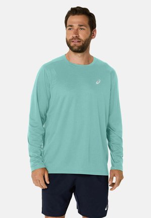 Chemise à manches longues turquoise clair, fabriquée dans un tissu lisse, col rond, arborant un petit logo sur la poitrine gauche. Portée avec un short bleu marine.