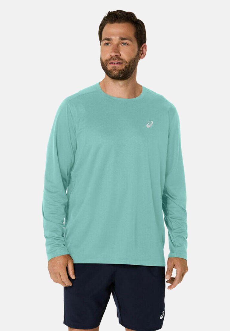 Camicia lunga azzurro chiaro, realizzata in tessuto morbido, colletto rotondo, con un piccolo logo sul lato sinistro del petto. Indossata con pantaloni corti navy.