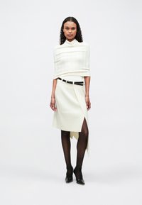 Donna con capelli ricci che indossa una gonna asimmetrica color crema, un top avvolgente coordinato, una cintura nera, collant neri trasparenti e tacchi neri, in piedi su uno sfondo bianco.