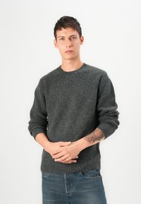 Dark gray wool sweater with ribbed texture and crew neckline, paired with blue denim jeans. Model stands with hands clasped.