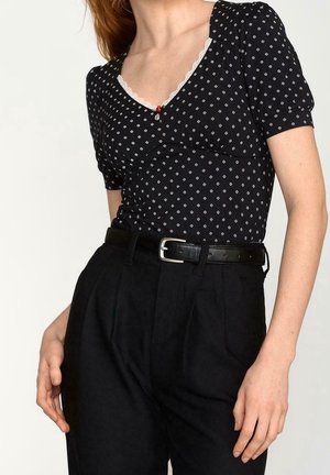 Kobieta w czarnej bluzce w grochy z krótkim rękawem i koronkową lamówką oraz czarnych spodniach z wysokim stanem i czarnym paskiem.