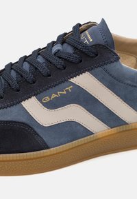 Zapatillas de ante y cuero en color azul marino, con acentos en beige y una suela de goma texturizada. Cordones y logo de GANT en detalle dorado.