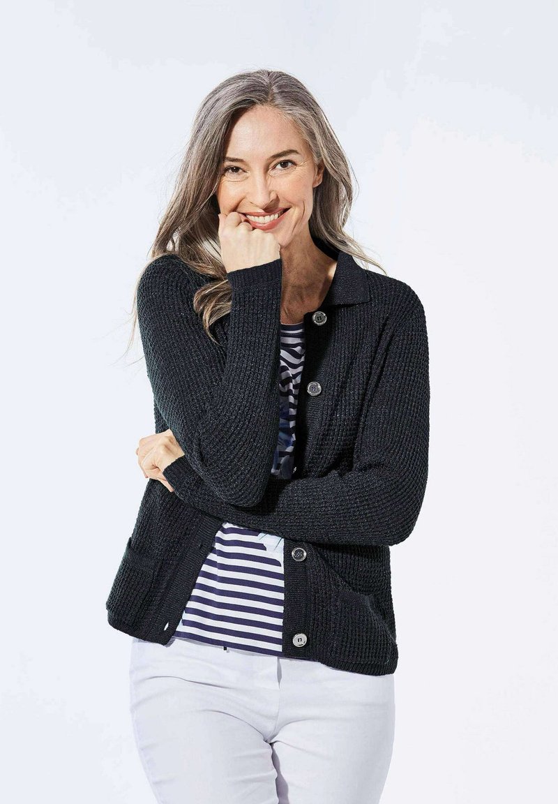 Schwarze Strickjacke mit Knopfverschluss und zwei aufgesetzten Taschen, getragen über einem gestreiften Hemd. Der Stoff hat eine strukturierte Oberfläche.