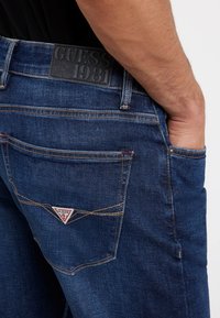 Tamsiai mėlyni džinsai su Guess logotipo aplikacija ant juosmens, kontrastinga siuvinėta ir trikampio logotipo detale ant nugarinės kišenės.