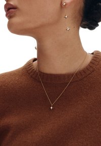 Zlatne naušnice u obliku zvijezde s malim bijelim detaljima, uz nježnu zlatnu ogrlicu s privjeskom zvijezde u istom stilu. Vidljiva struktura smeđeg džempera.