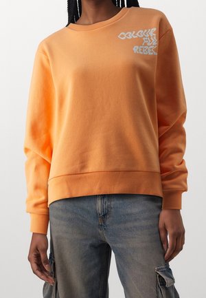 Persoon draagt een oranje sweatshirt met de tekst "Colour Full Rebel" bij de schouder, gecombineerd met blauwe cargobroek.