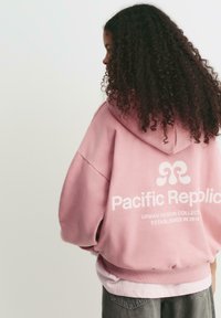 Person med långt lockigt hår iklädd en rosa hoodie med texten "Pacific Republic Urban Design Collection Established 2018" på ryggen.