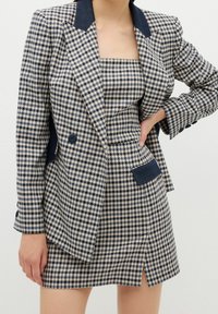 Femme portant un blazer pied-de-poule bleu marine et beige avec un top sans manches et une jupe assortis, avec un col bleu marine foncé et des détails de poche.