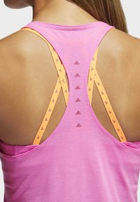 Gros plan sur le haut du dos d'une femme portant un débardeur rose par-dessus un soutien-gorge de sport orange avec plusieurs logos adidas sur les bretelles.