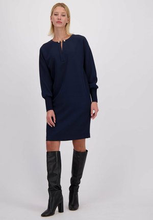 Marineblauwe jurk met een ronde hals en knoopdetails, lange mouwen en een relaxte pasvorm. Gecombineerd met kniehoge zwarte leren laarzen.