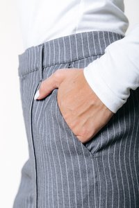 Χέρι με λευκό βερνίκι νυχιών που ξεκουράζεται μέσα στην τσέπη γκρίζων παντελονιών με ρίγες, φορεμένο με ένα άσπρο μπλουζάκι με μακρύ μανίκι.