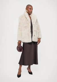 Kreemjas valejõhvi jope laia kraega, pikkade varrukatega ja kahe eesmise taskuga, kantud tumeda pruunist satiinkleidi üle ning hoides lilat käekotti.