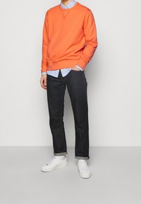 Orange sweatshirt med rund hals, bärs över en ljusblå mönstrad skjorta; ihop med mörka jeans och vita sneakers. Grönt logotypdetalj synlig.