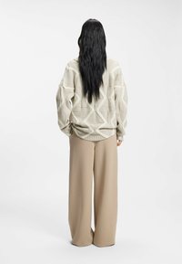 Jersey beige de punto con patrón de diamantes texturizado, combinado con pantalones anchos beige. Visto desde atrás, cabello largo y oscuro cayendo hacia abajo.