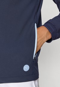 Navy jacket with a textured fabric, light blue zip, elastic cuffs, and a circular light blue logo on the hem, highlighting a sports design.