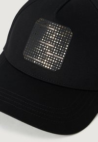 Melna auduma cepure ar teksturētu zelta logo plati, uz kuras ir uzraksts "Armani Exchange" un punktiņu raksti priekšpusē. Izstrādāta ar izliekto malu.