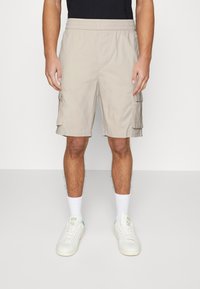 Hombre vestido con pantalones cortos tipo cargo beige hasta la rodilla, calcetines blancos y zapatillas blancas, de pie contra un fondo liso.