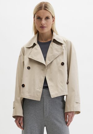 Woman wearing a cropped beige trench coat with dark buttons over a dark ribbed shirt and gray pants, standing against a white background.