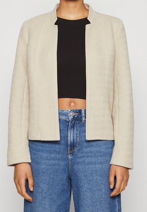 Personne portant une veste beige ouverte, un haut court noir et un jean bleu taille haute, debout les bras détendus le long du corps.