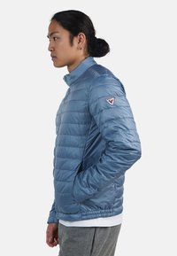 Blå puffjacka med horisontellt quiltade sektioner, hög krage och sidofickor. Har en logotyp-patch på vänster axel.