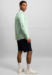 Camisa de botón a rayas verdes y blancas con mangas largas, combinada con pantalones cortos azul marino y zapatillas blancas. Tela suave, corte relajado.