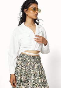 Camisa branca cropped de mangas longas com um bolso, combinada com uma saia preta floral com detalhes em tons pastel e uma cintura elástica.
