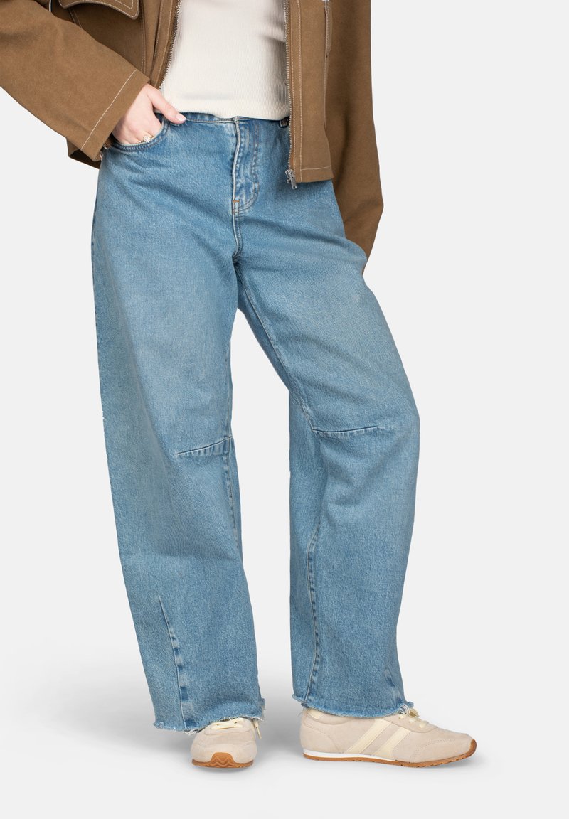 Personne portant un jean bleu clair à jambes larges avec des fentes à l'avant, des baskets beige, un haut blanc et une veste marron les mains dans les poches.