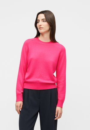 Vrouw met recht bruin haar, gekleed in een felroze trui met ronde hals en zwarte broek, staand tegen een effen lichte achtergrond.