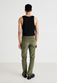 Man standing facing away wearing black sleeveless top, olive green pants, and black shoes on plain light background.