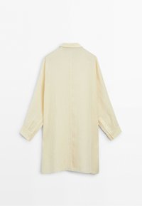 Long cream-colored shirt dress with long sleeves, buttoned cuffs, and a simple collar, shown from the back on a white background.