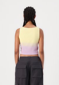 Vrouw met gevlochten haar, gekleed in een mouwloze crop top met een geel naar lila verloop en donkere cargobroeken, die met haar rug naar de camera staat tegen een witte achtergrond.