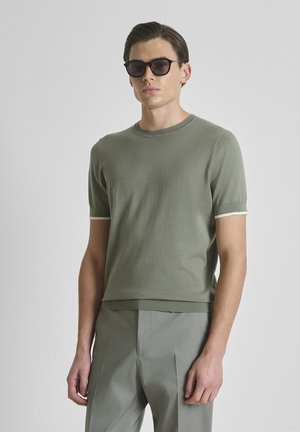 Uomo con capelli castani corti che indossa occhiali da sole neri, maglione a maniche corte color oliva chiaro e pantaloni grigi, in piedi davanti a uno sfondo semplice.